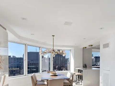 A white dining area with corner windows onto the city, a branching chandelier and glass-front wall cabinets