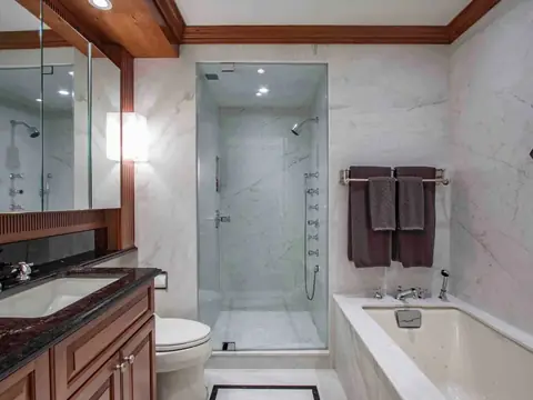 A bathroom with white marble walls, a glass-enclosed shower, a soaking tub in a marble deck and a cherry vanity with a black granite top