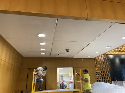 Two workers on rolling scaffolds at the ceiling of a wood-panelled room with new ceiling panels and recessed lights