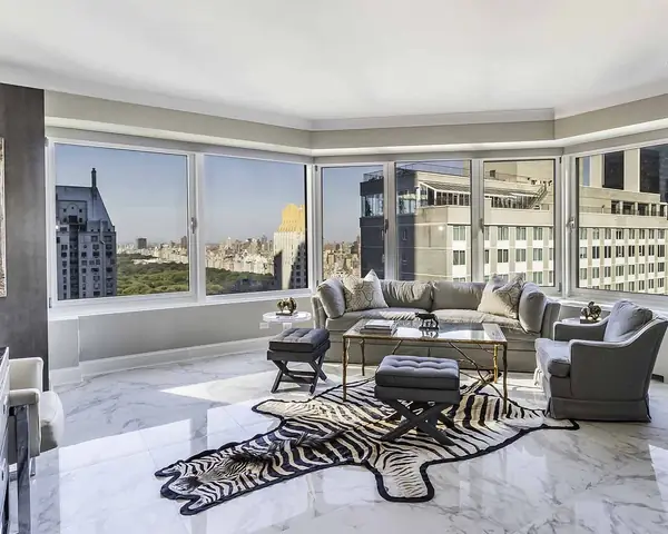 A living room with corner windows onto a park and city towers, grey sofas and a zebra hide rug on a white marble floor