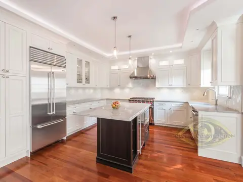 A kitchen with white cabinets to the ceiling, a dark island with a white top, a steel refrigerator, two pendants and a cherry floor