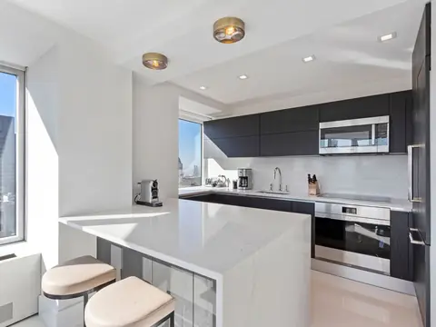 A white kitchen with a waterfall peninsula, dark wall cabinets, a built-in oven and microwave and windows onto city towers